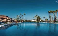 Hotelpool mit Liegestühlen und Palmen unter blauem Himmel.
