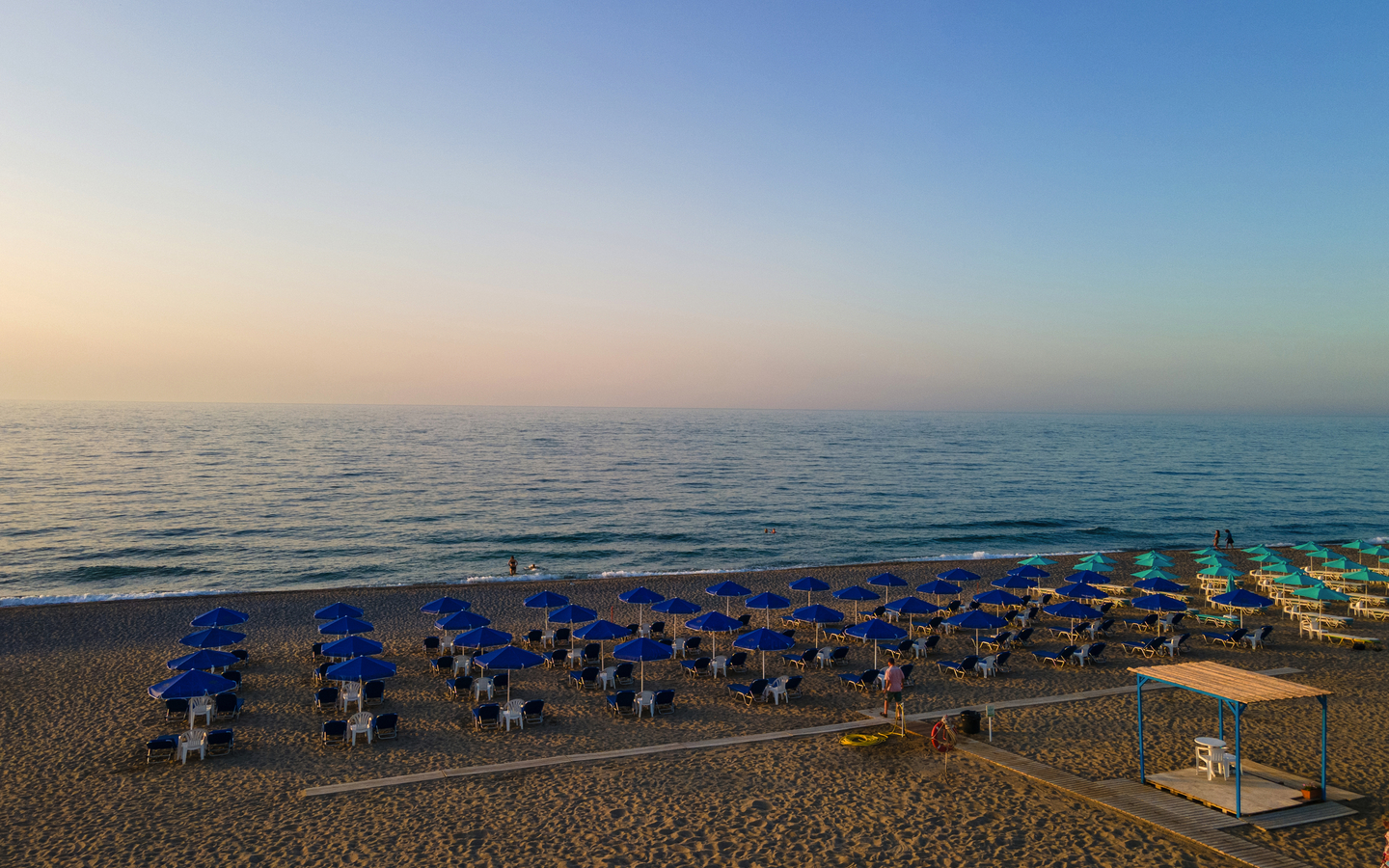 Strand mit vielen blauen Sonnenschirmen und klarem Meer im Hintergrund.