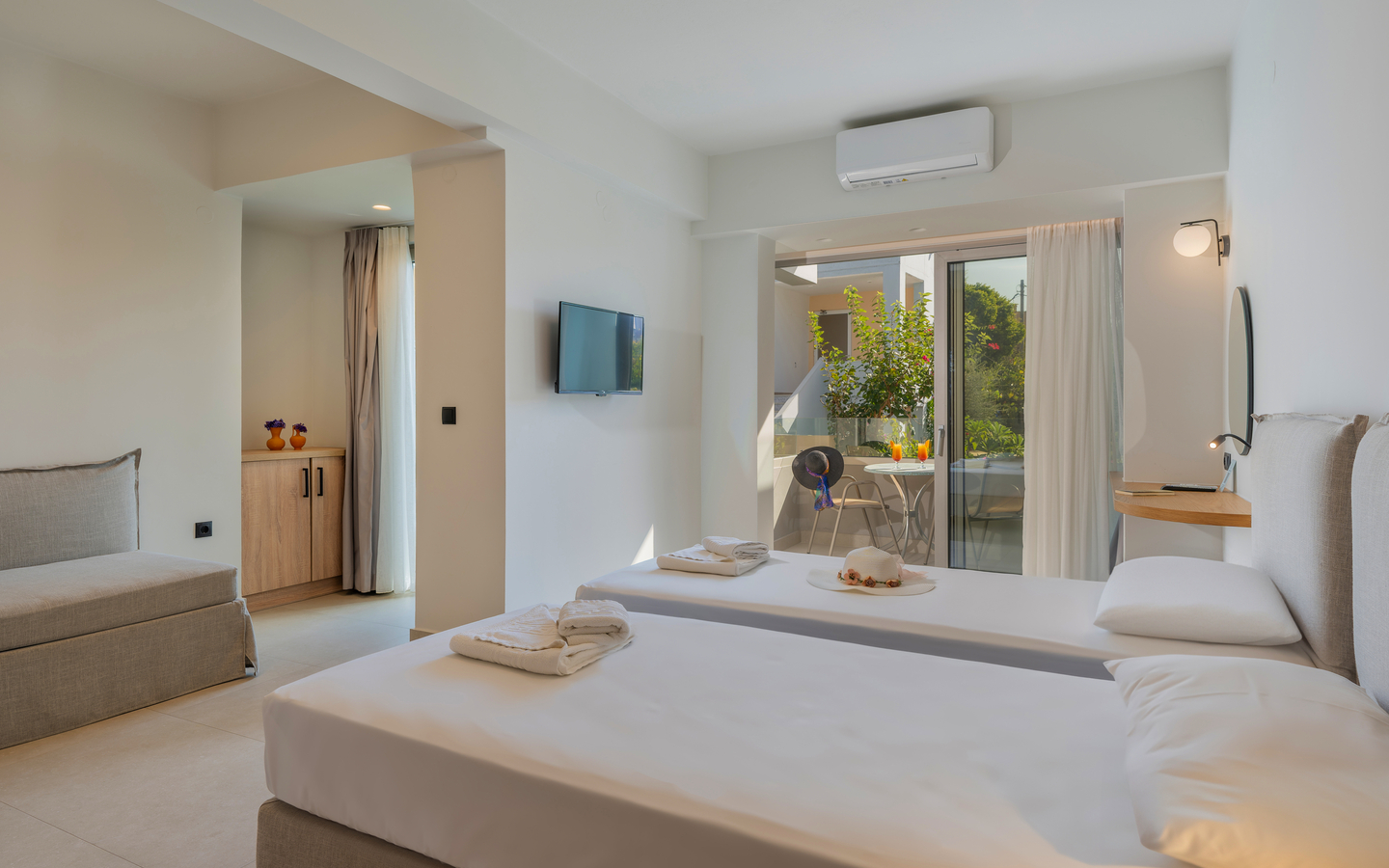 Helles Hotelzimmer mit Doppelbett, Sofa, TV und Balkon mit Ausblick auf Grünfläche.
