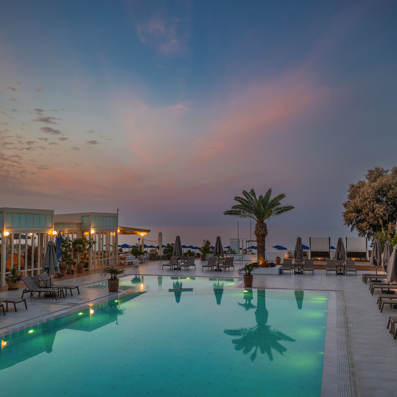 Hotelpool bei Sonnenuntergang mit Liegestühlen und Palmen in Griechenland.