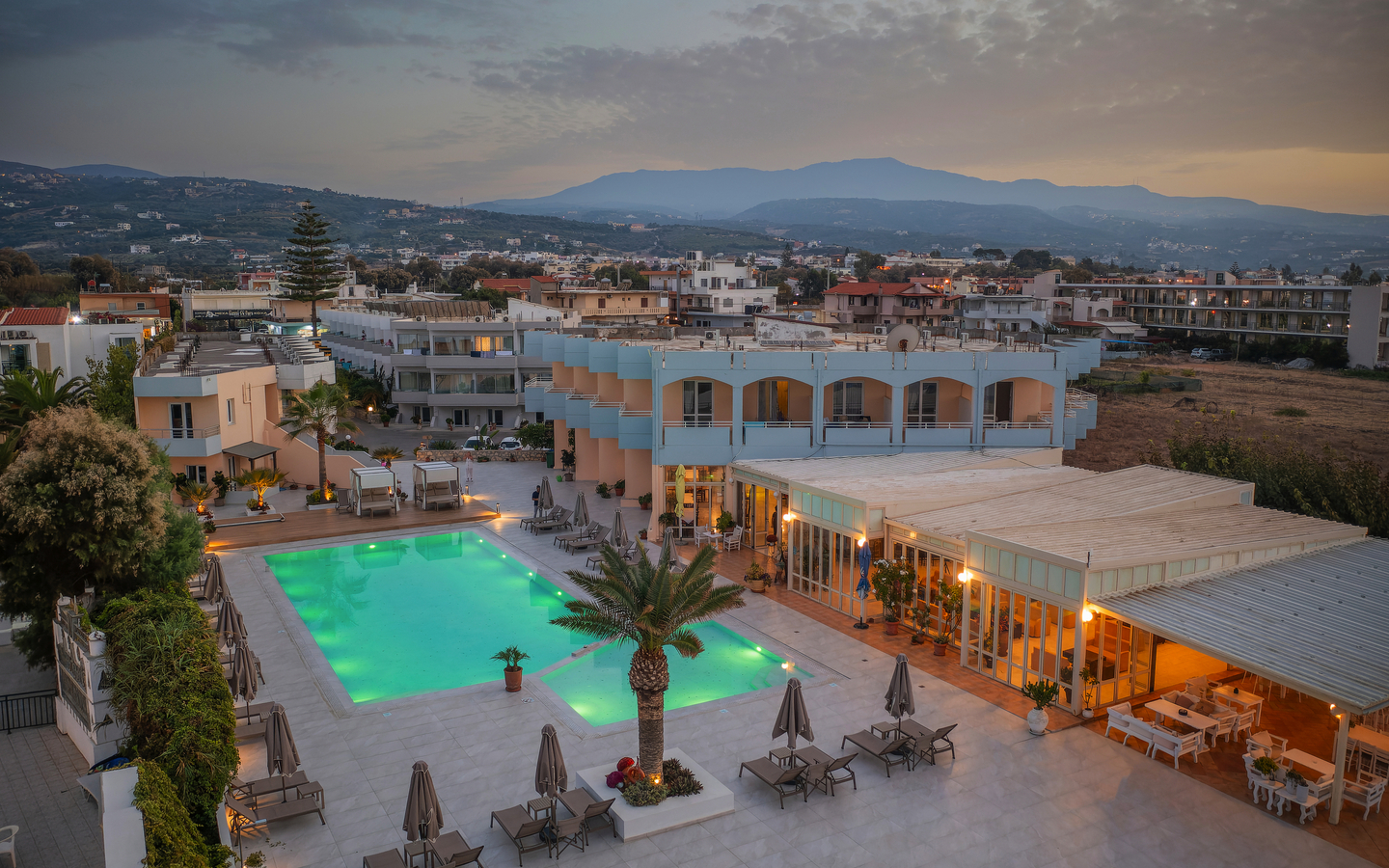 Hotel mit Pool auf Kreta bei Sonnenuntergang, umgeben von Gebäuden und Hügeln.