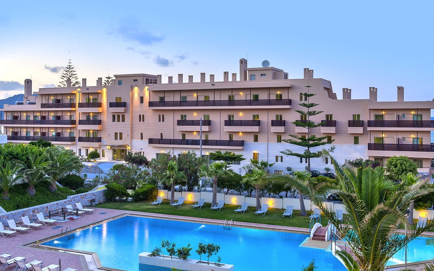 Hotel mit Pool, umgeben von Palmen, nahe einem Strand bei Sonnenuntergang.