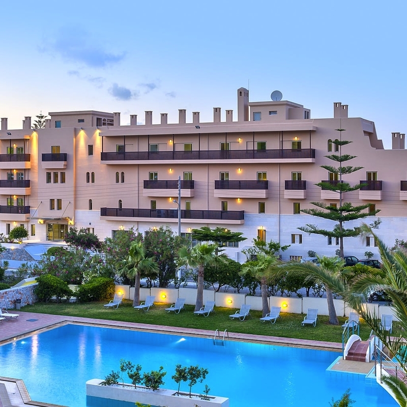 Hotel mit Pool, umgeben von Palmen, nahe einem Strand bei Sonnenuntergang.