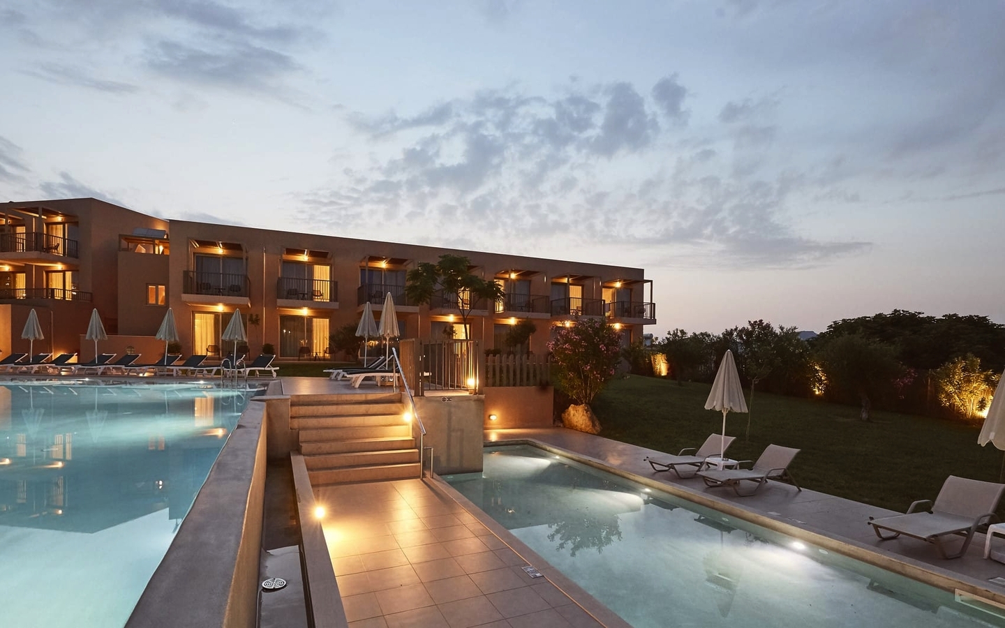 Außenansicht eines Hotels mit Pool und Liegestühlen bei Sonnenuntergang.