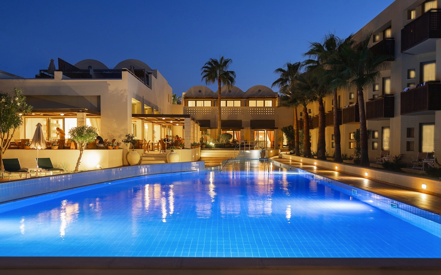 Außenansicht des beleuchteten Hotelpools am Abend, umgeben von Palmen.