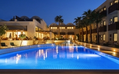 Außenansicht des beleuchteten Hotelpools am Abend, umgeben von Palmen.