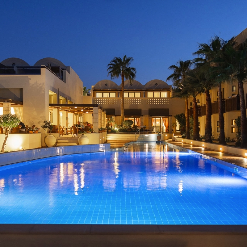 Außenansicht des beleuchteten Hotelpools am Abend, umgeben von Palmen.