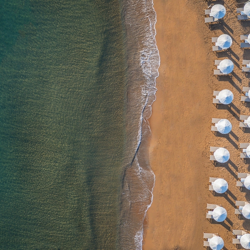 Strand mit Sonnenschirmen und Wellen am Ufer aus der Vogelperspektive.