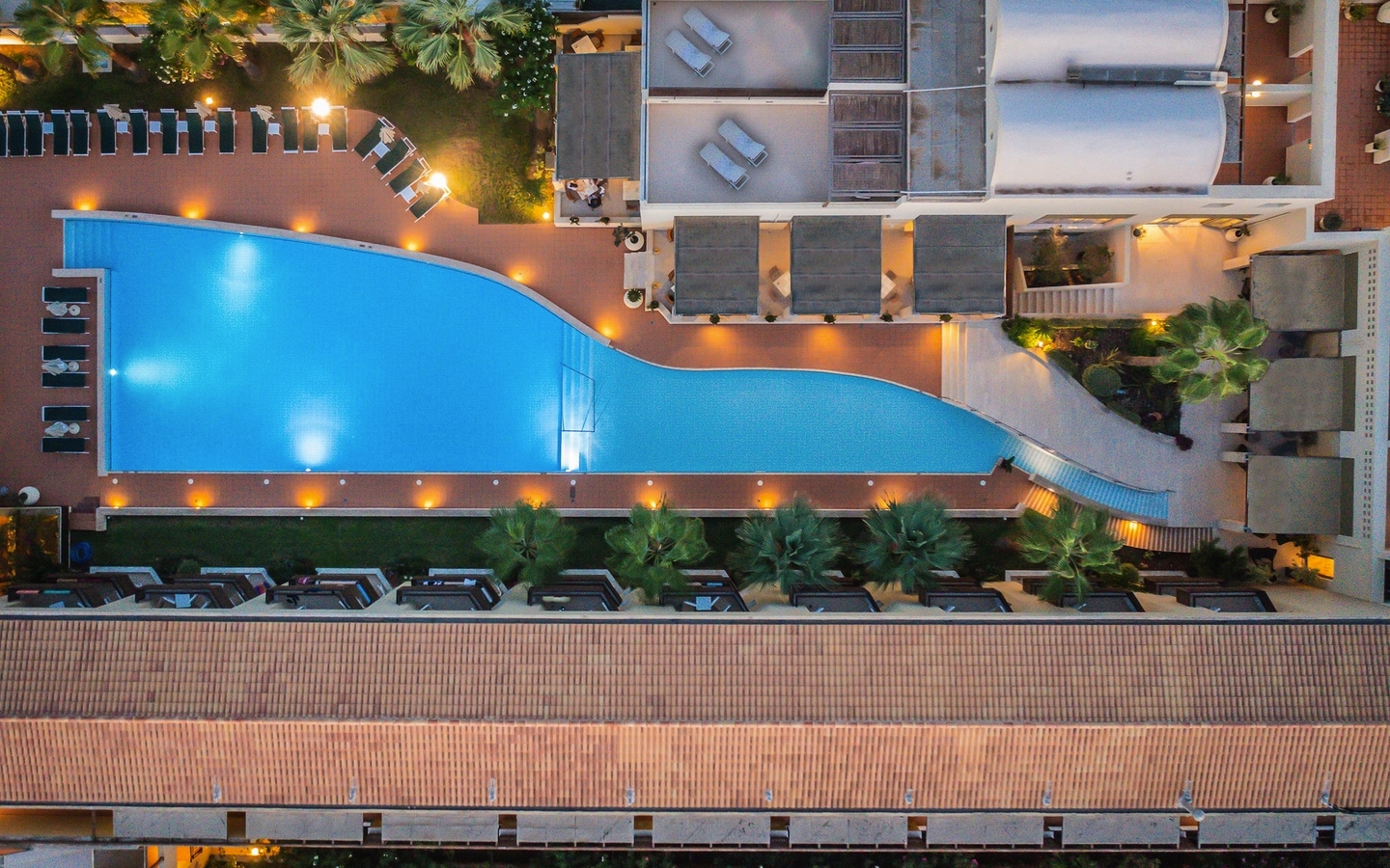 Hotelpool und Sonnenliegen von oben, umgeben von Palmen, am Abend beleuchtet.