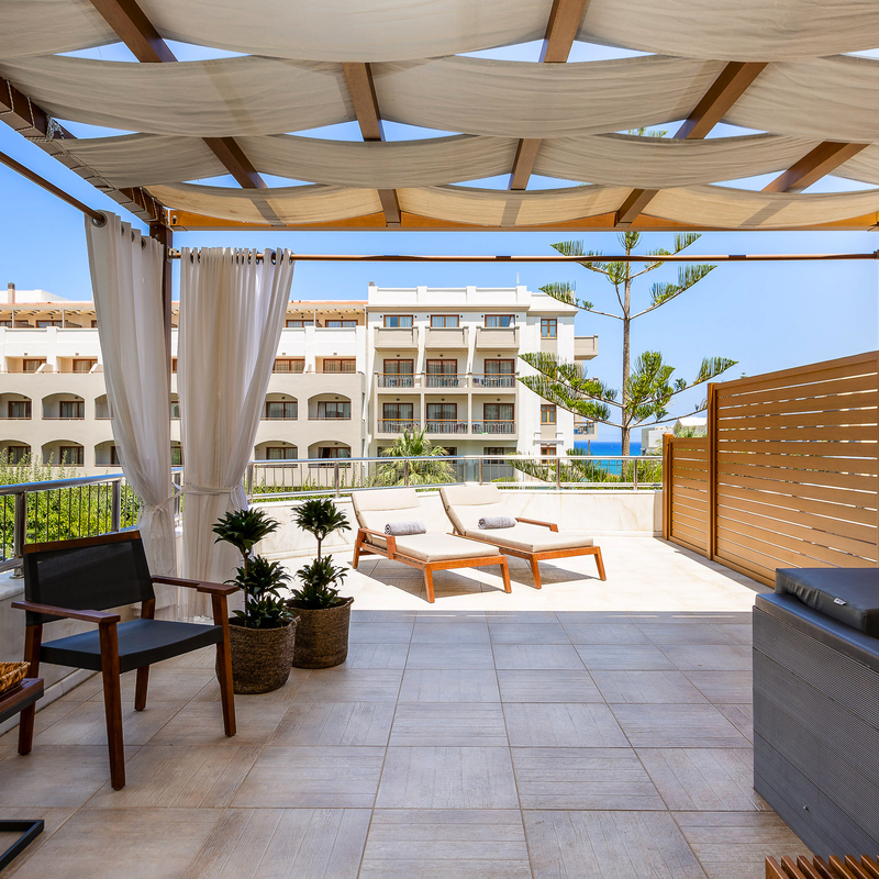 Terrasse einer Suite mit Jacuzzi, Sonnenliegen und Blick auf Gebäude im Hintergrund.