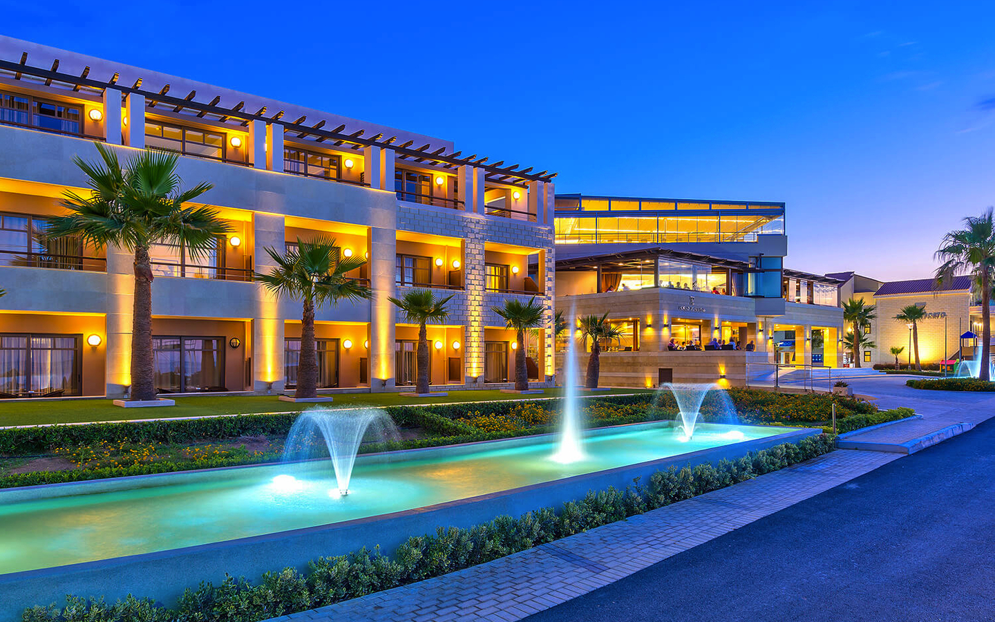 Außenansicht eines modernen Hotels mit Palmen und beleuchteten Wasserfontänen bei Abenddämmerung.