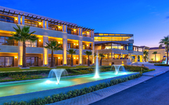 Außenansicht eines modernen Hotels mit Palmen und beleuchteten Wasserfontänen bei Abenddämmerung.