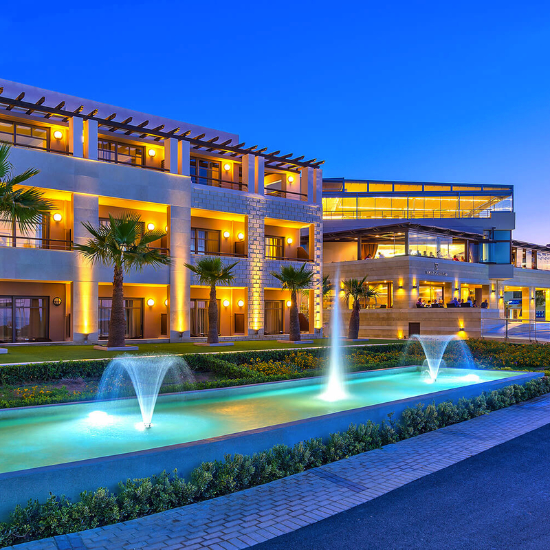 Außenansicht eines modernen Hotels mit Palmen und beleuchteten Wasserfontänen bei Abenddämmerung.