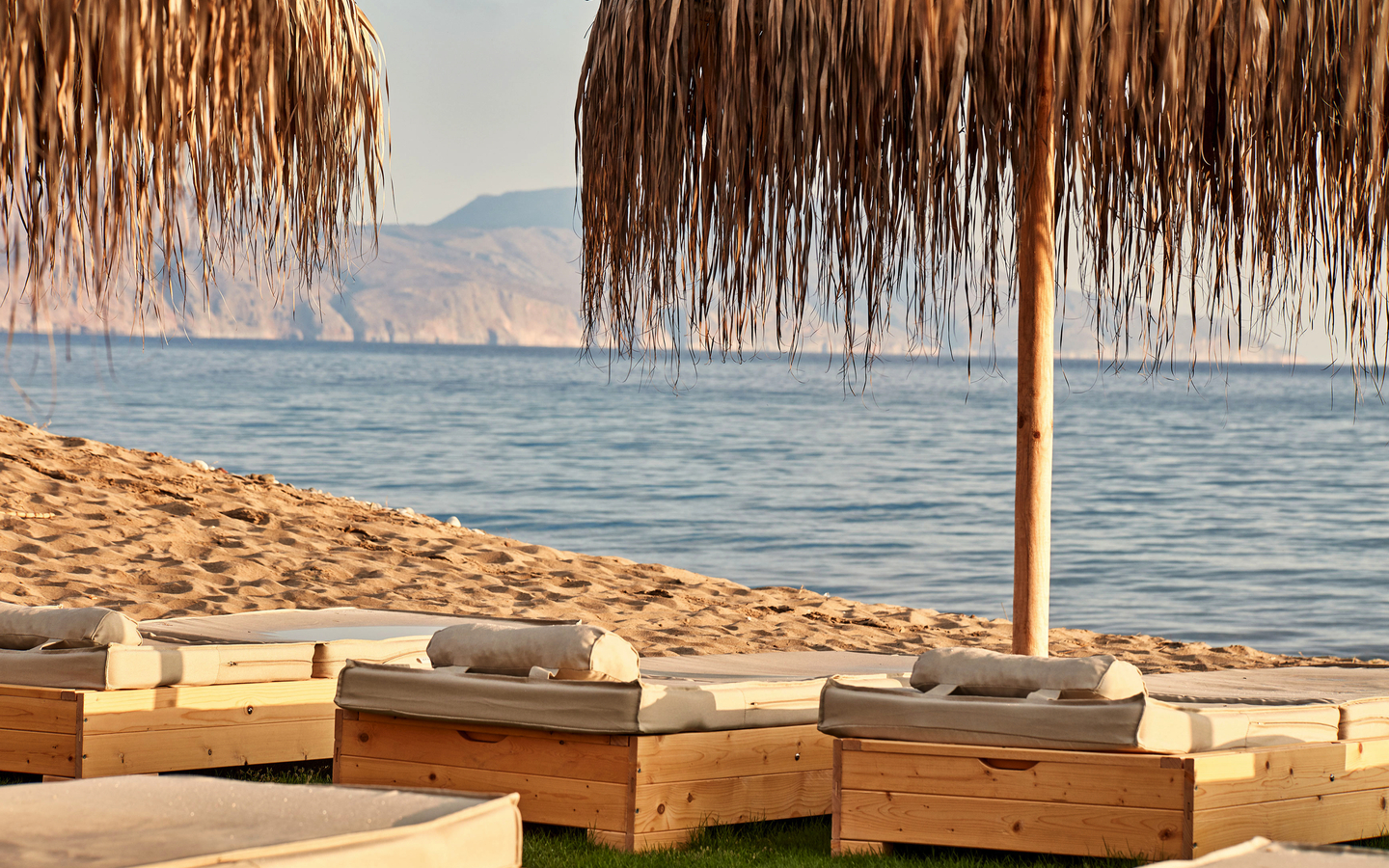Strand mit Liegen unter Strohdächern, im Hintergrund das Meer und Hügel.