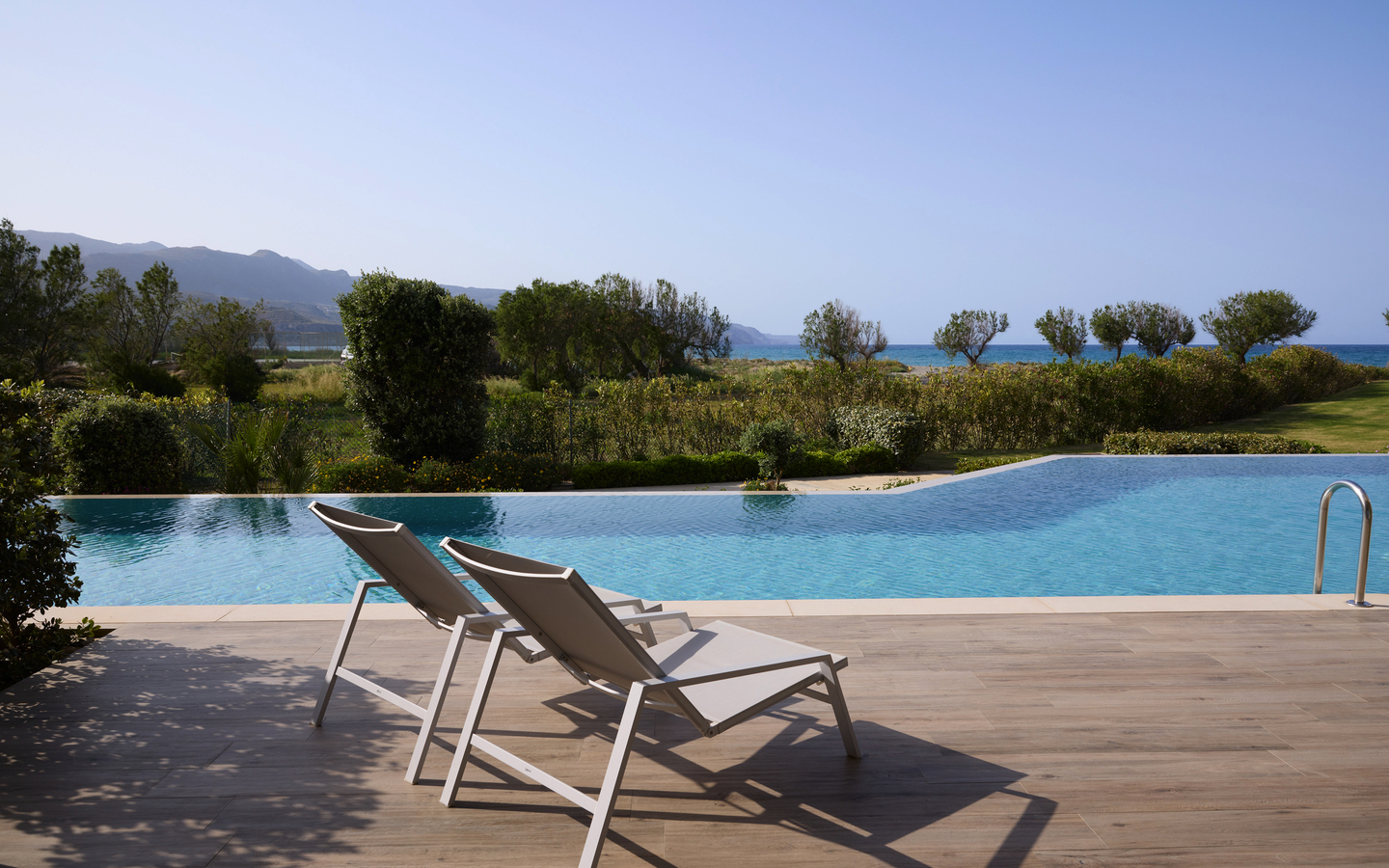 Zwei Liegestühle auf einer Terrasse mit Blick auf einen Pool und das Meer im Hintergrund.