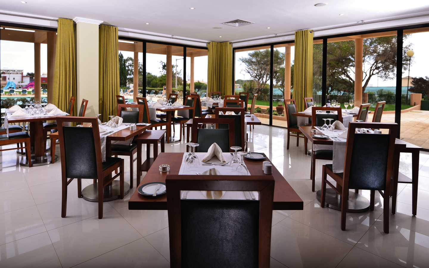 Helles Restaurant mit gedeckten Tischen und großen Fenstern, die in einen Garten blicken.