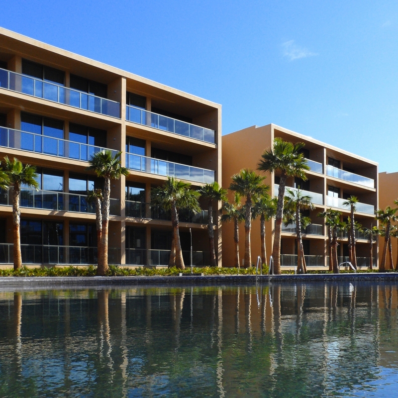 Hotelgebäude mit Palmen am Pool, blauer Himmel im Hintergrund.