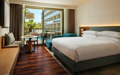 Hotelzimmer mit Doppelbett, Sofa, Tisch und Balkon mit Blick auf Palmen.