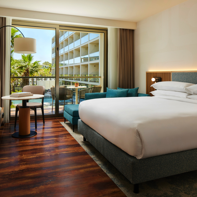 Hotelzimmer mit Doppelbett, Sofa, Tisch und Balkon mit Blick auf Palmen.