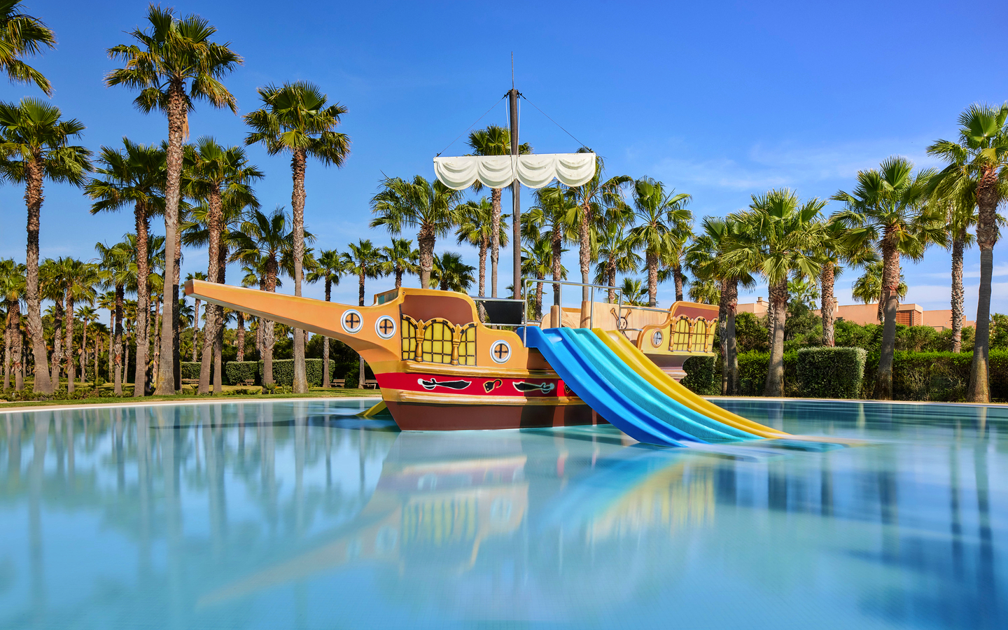 Spielplatz im Wasser mit Piratenschiff und Rutschen, umgeben von Palmen, unter blauem Himmel.