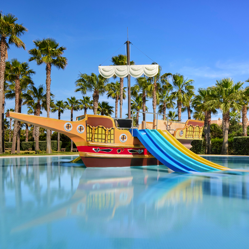 Spielplatz im Wasser mit Piratenschiff und Rutschen, umgeben von Palmen, unter blauem Himmel.