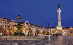 Springbrunnen und Statue auf einem Platz bei Nacht in einer europäischen Stadt.