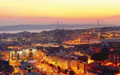 Städtische Skyline bei Sonnenuntergang, beleuchtete Gebäude und Brücke im Hintergrund.