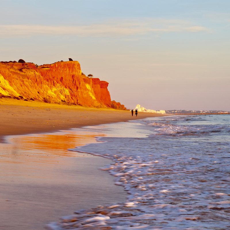 Küste mit roten Klippen und zwei Personen bei Sonnenuntergang am Strand.