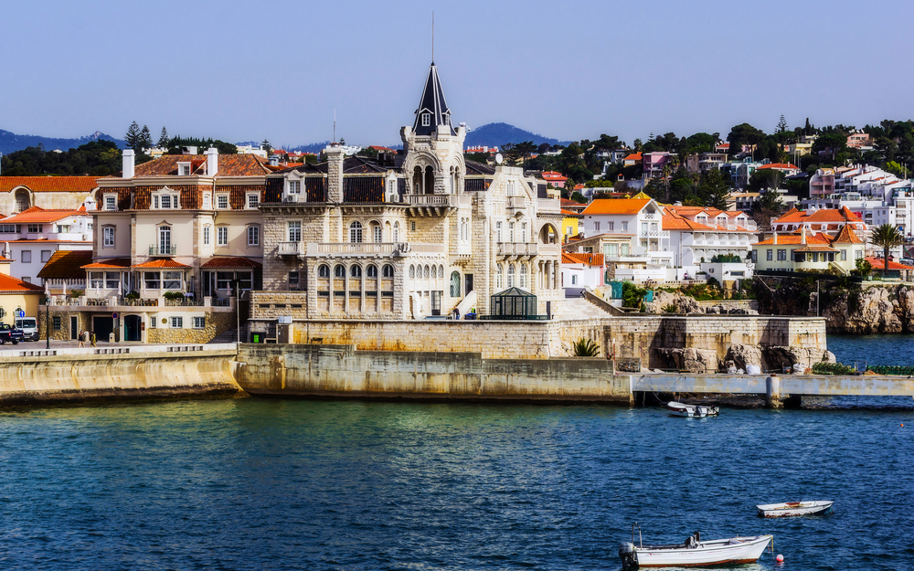 Küstenlandschaft in Estoril, Portugal mit historischem Gebäude und Booten im Wasser.