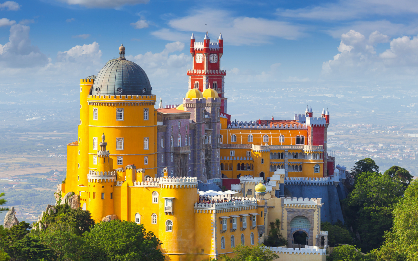Gelbes Schloss auf einem Hügel, umgeben von Wald, mit blauem Himmel im Hintergrund.