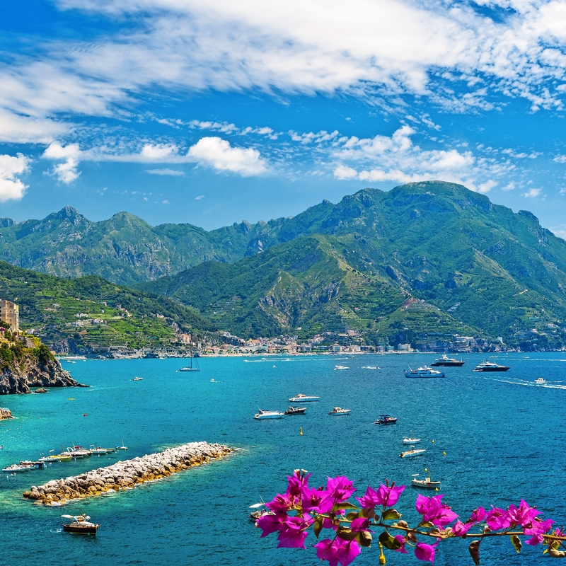 Küstenstadt mit bunten Häusern und Booten am blauen Meer, Bergen im Hintergrund.