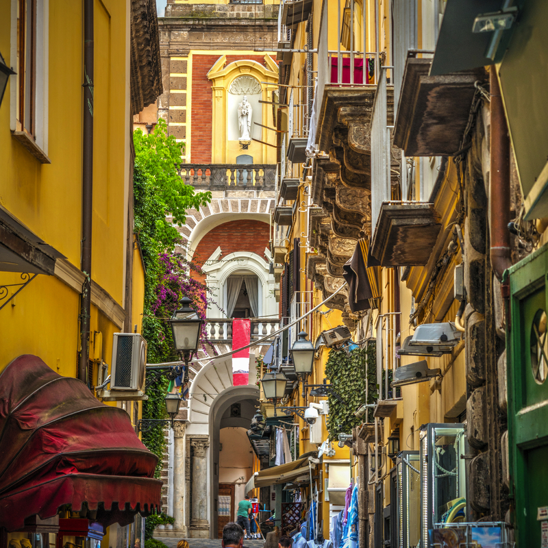 Schmale, belebte Gasse mit gelben Gebäuden und Balkonen in einer historischen Stadt.