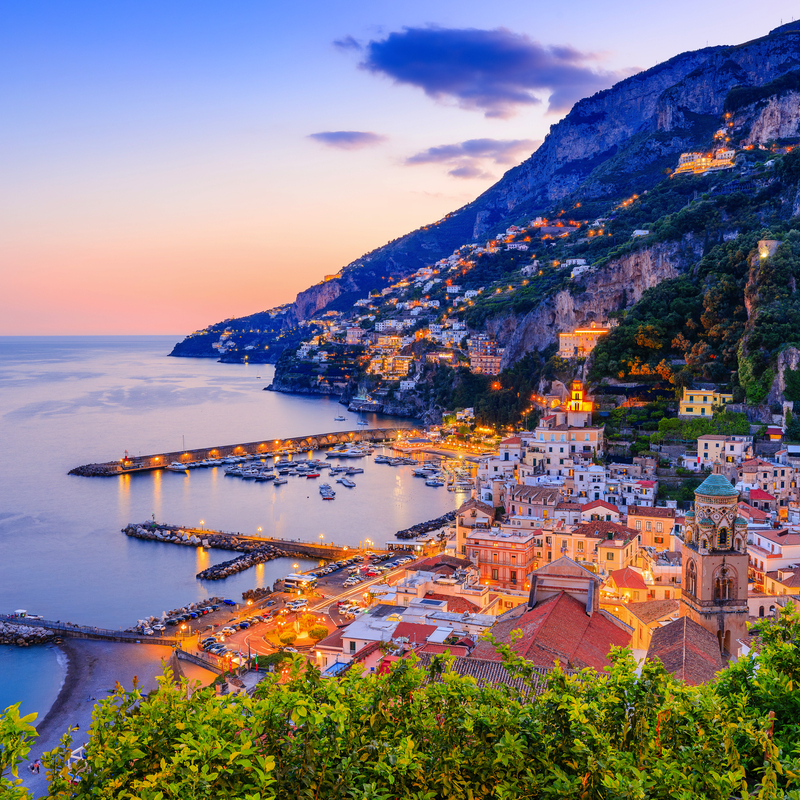 Küstenstadt bei Sonnenuntergang mit beleuchteten Häusern und Hafen am Meer.