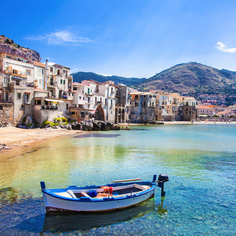 Boot auf klarem Wasser vor Küste mit Häusern und Bergen im Hintergrund bei Sonnenschein