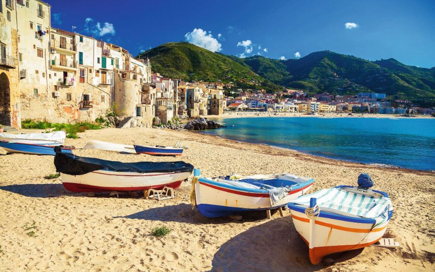 Boote liegen am Strand in einer mediterranen Küstenstadt vor grünen Hügeln und blauem Meer.
