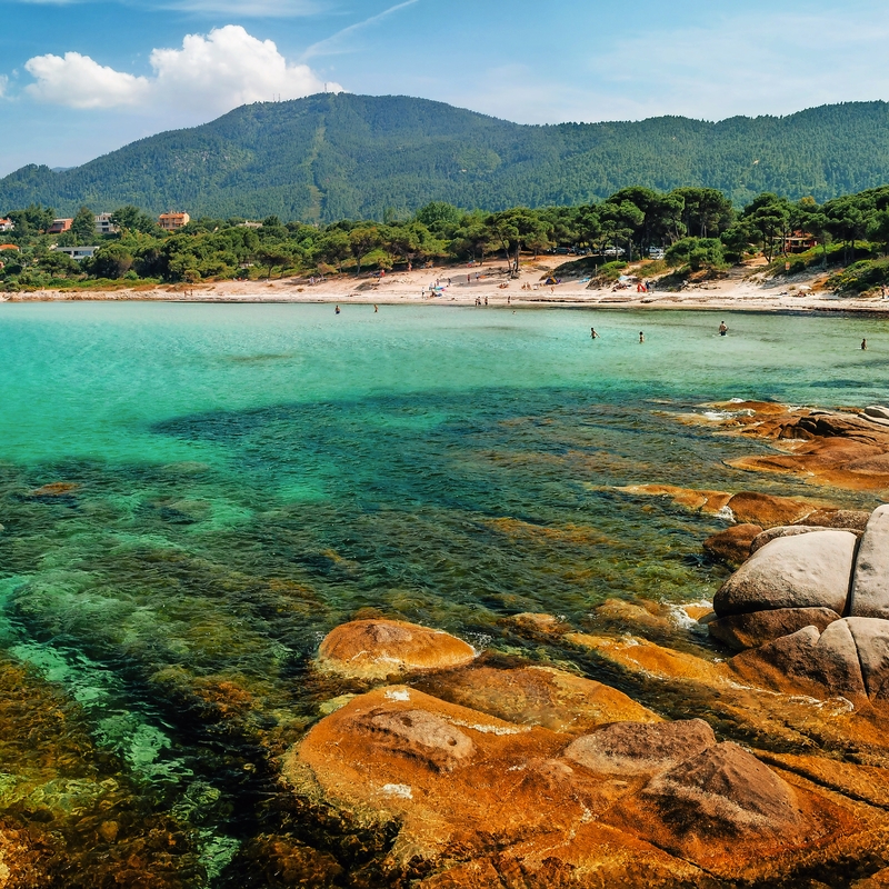 Küstenlandschaft mit klarem Wasser, Felsen und bewaldeten Hügeln im Hintergrund.