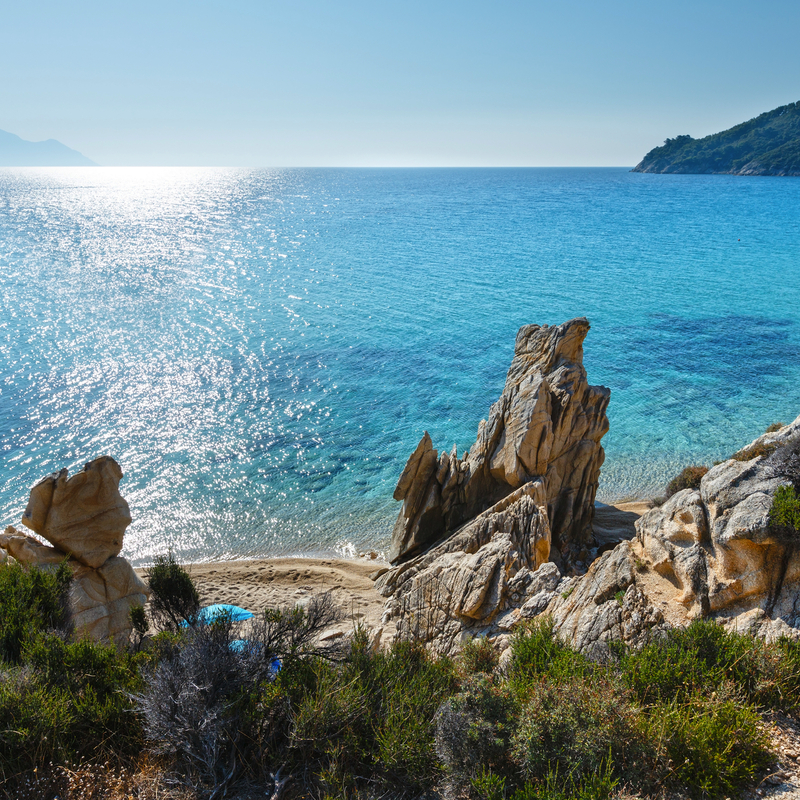 Küste mit Felsen und kristallklarem Wasser unter blauem Himmel.