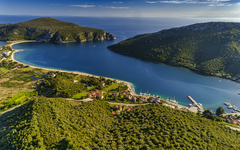 Bucht mit blauem Wasser und grünen Hügeln, kleine Gebäude und Boote am Ufer.