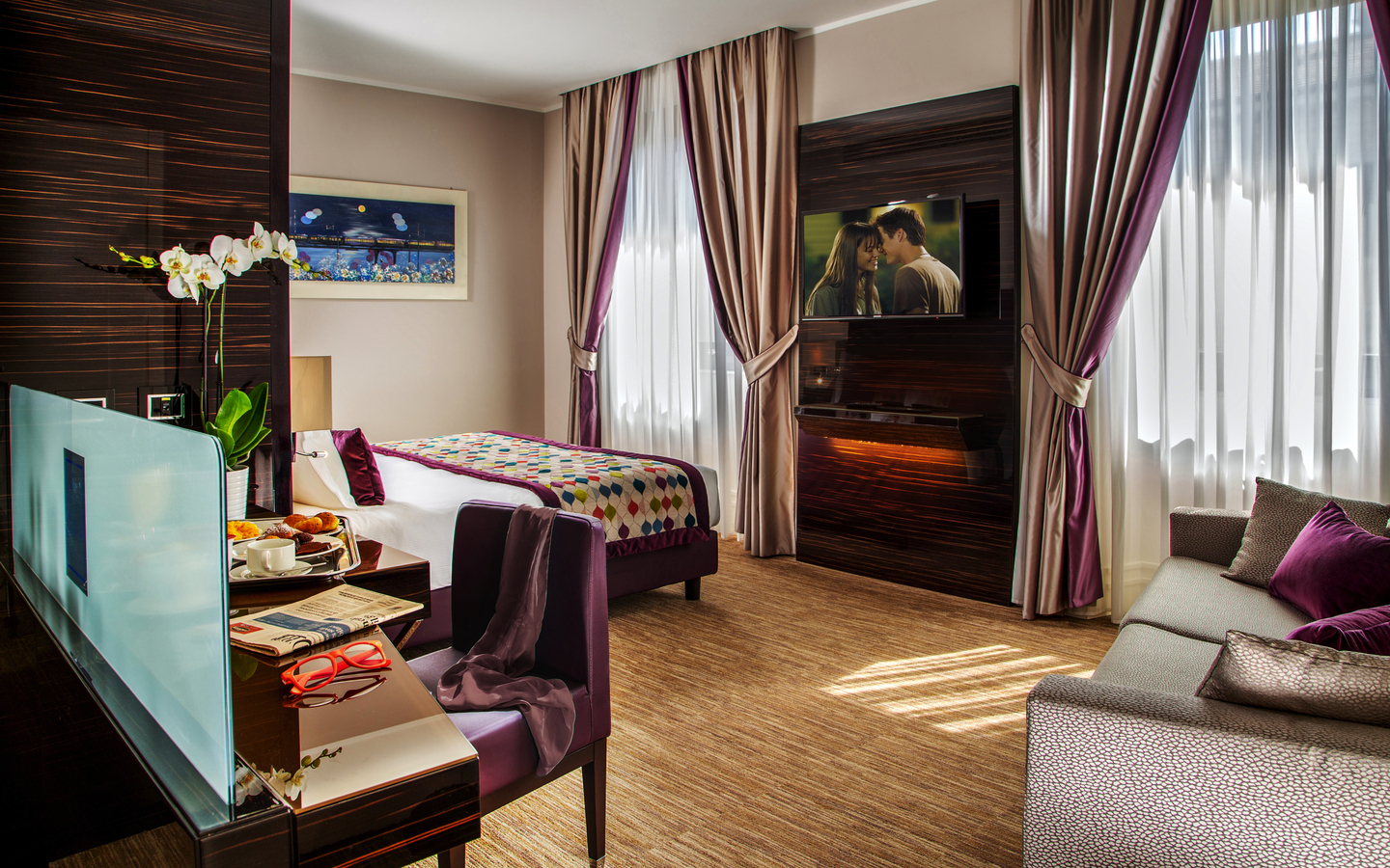 Modern eingerichtetes Hotelzimmer mit Doppelbett, Sofa, Fernseher und Frühstückstablett.