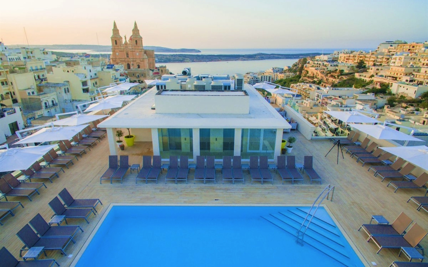 Terrasse mit Pool und Liegestühlen, im Hintergrund Meerblick und Gebäude.