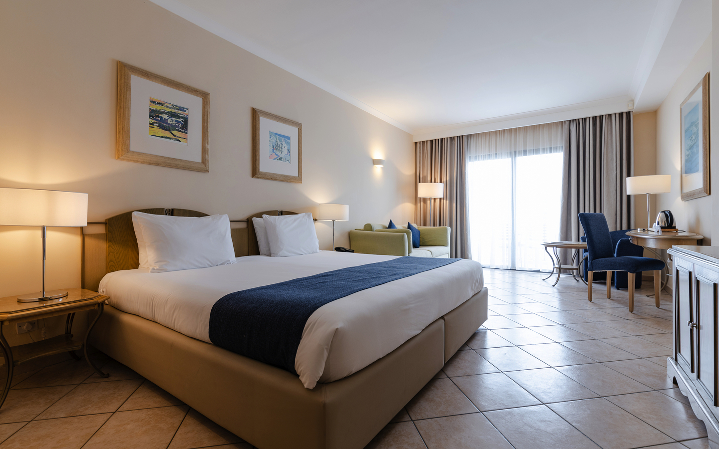 Helles Hotelzimmer mit Doppelbett, Sofa, Tisch und großen Fenstern mit Vorhängen.