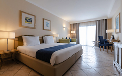 Helles Hotelzimmer mit Doppelbett, Sofa, Tisch und großen Fenstern mit Vorhängen.