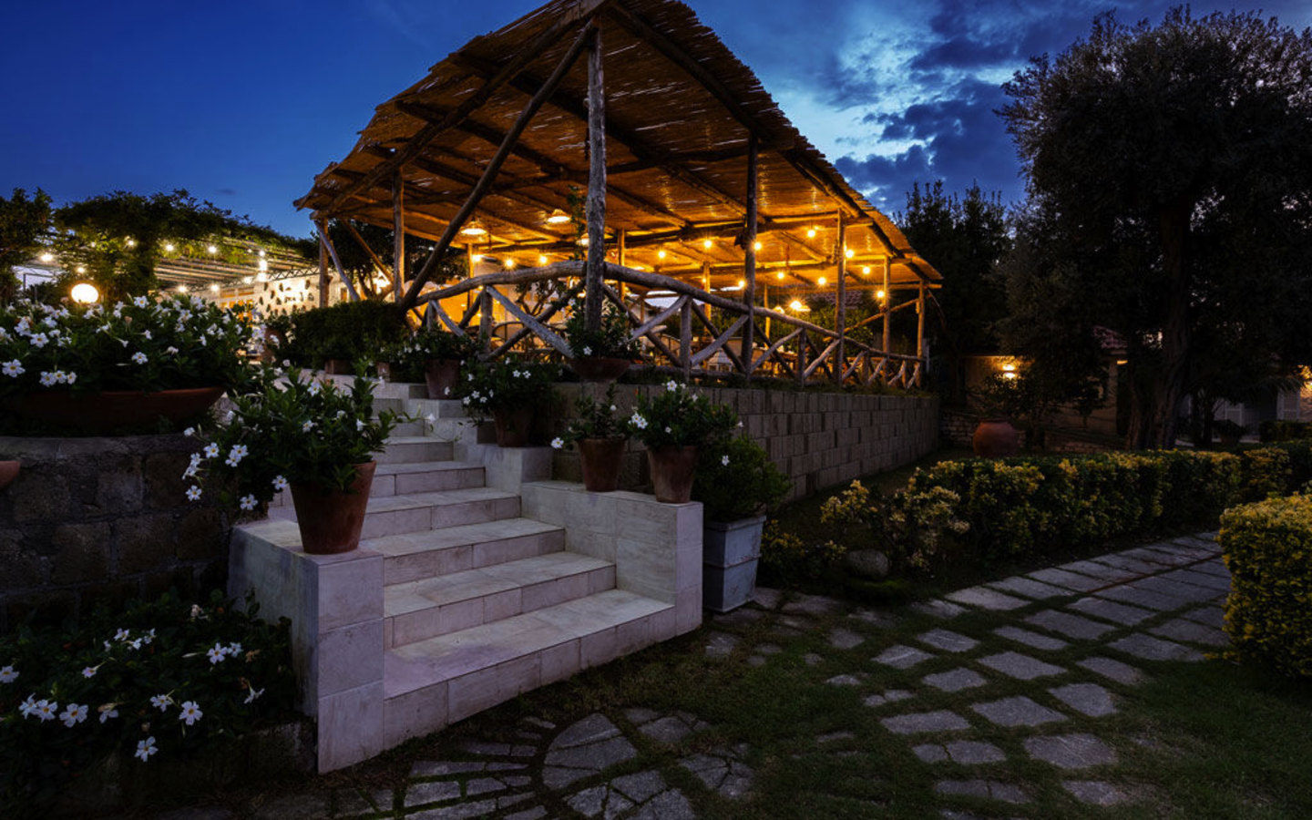 Beleuchtete Terrasse eines Restaurants am Abend mit blühenden Pflanzen und Steinweg.