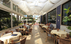 Hotelterrasse mit Gästen an Tischen, Sonnenschirmüberdachung und Blick auf Garten.
