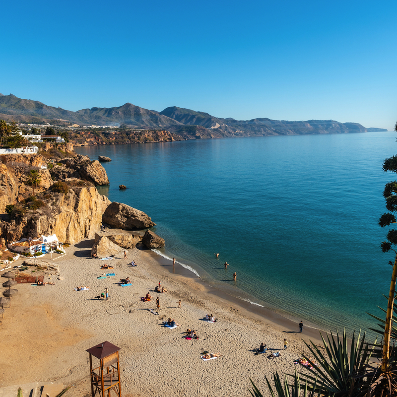 Strand in Nerja, Costa del Sol, mit Sand, Felsen, Meer und Bergen im Hintergrund.