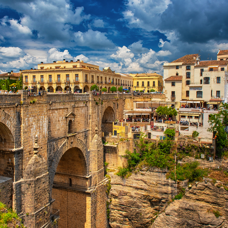 Historische Steinbrücke in Ronda, umgeben von Gebäuden und bewölktem Himmel.