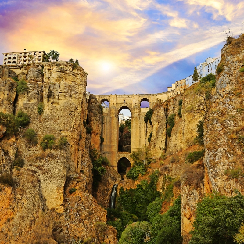 Steinbrücke über eine tiefe Schlucht bei Sonnenuntergang mit Gebäuden am Rand.