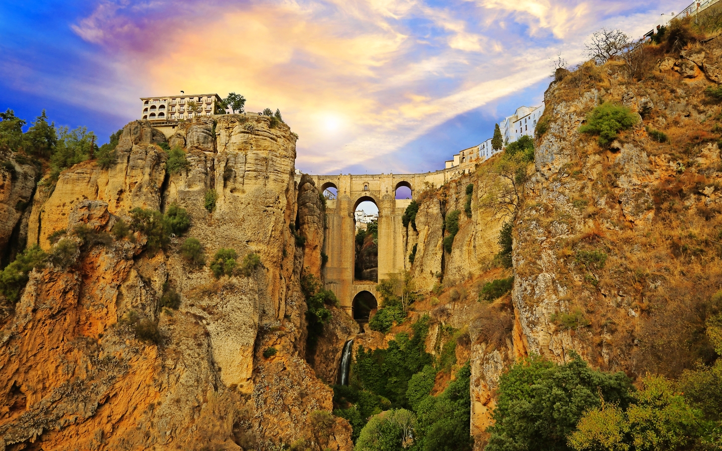 Steinbrücke über eine tiefe Schlucht bei Sonnenuntergang mit Gebäuden am Rand.