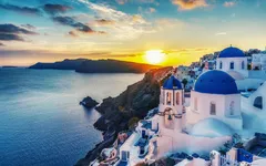 Panoramablick auf Oia, Santorin bei Sonnenuntergang mit blau-weißer Kirche im Vordergrund.