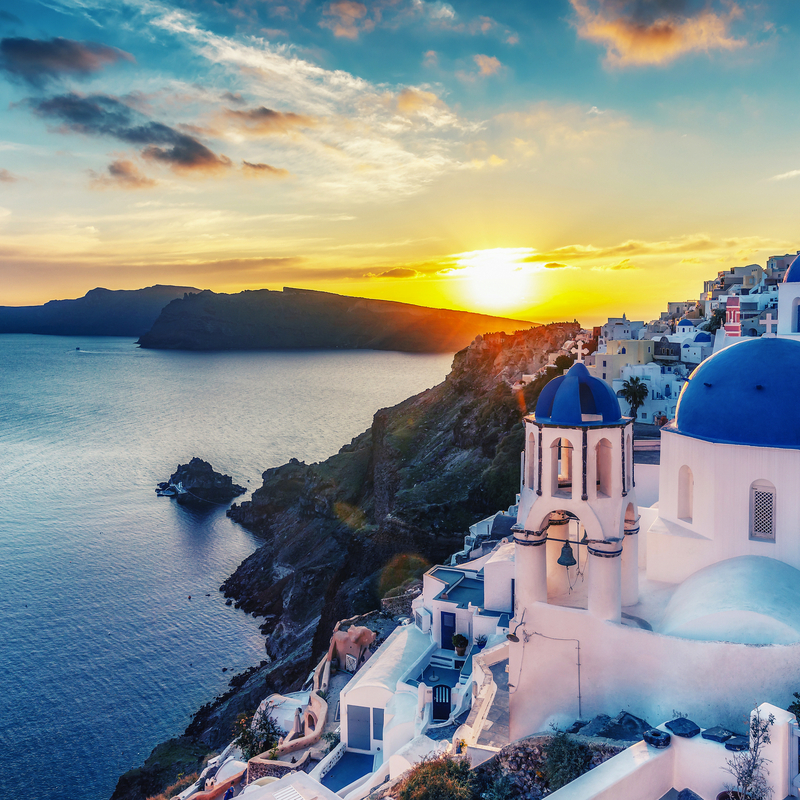 Panoramablick auf Oia, Santorin bei Sonnenuntergang mit blau-weißer Kirche im Vordergrund.
